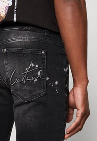 Mustat farkut kapealla istuvuudella, joissa on takataskusomiste, valkoinen roiskemainen kuvio ja logopistely taskussa.