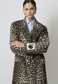 Femme aux cheveux plaqués en arrière portant un blazer oversized à imprimé léopard sur une chemise bleu clair, avec un patch de cygne noir sur la manche, bras croisés.