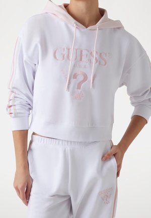 Person iført hvid cropped hættetrøje og matchende sweatpants med lyserødt "GUESS"-logo og spørgsmålstegnsdesign, hånd i lommen.