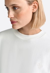 Sudadera blanca con cuello redondo acanalado y un logo texturizado en el pecho. Tejido suave con un acabado blando.