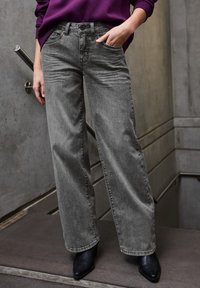 Graue weit geschnittene Jeans aus Denim mit hohem Bund, vorderen Taschen und subtiler Textur. Kombiniert mit schwarzen Absatzstiefeln.