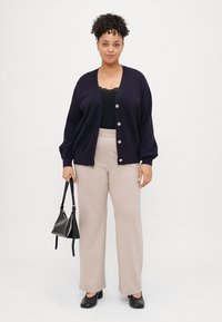 Cardigã azul-marinho com seis botões prateados, combinado com uma blusa preta e calças largas bege. Bolsa preta e sapatos pretos.