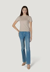 Camiseta de algodón beige con el logo "GUESS" en acabado brillante, combinada con vaqueros acampanados azul claro y zapatos de tacón beige.