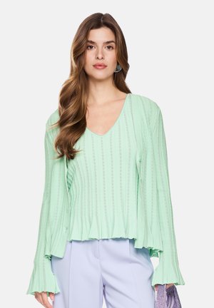 Woman with long wavy brown hair wearing a mint green textured V-neck sweater and light lavender pants, holding a purple knit accessory.