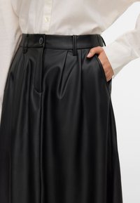 Pantalons en simili cuir noir avec plis, maintenus par une taille et une fermeture à bouton. Poches latérales visibles. Texture lisse.