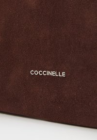 Braunes Wildledermaterial mit einer weichen Textur, das ein silbernes, auf die Oberfläche geprägtes "COCCINELLE"-Logo zeigt. Kompakte und rechteckige Form.