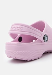 Pink Crocs with a rounded shape, made of textured material. Features a back strap and a circular logo accent. Patterned sole for traction.