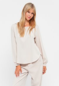 Μπλούζα μπεζ με μακριά μανίκια και στρογγυλή λαιμόκοψη, απαλή υφή και ρυτιδωτά μανίκια, σε συνδυασμό με αντίστοιχες ανοιχτόχρωμες παντελόνες.