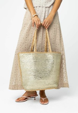 Žena v béžových šatách s očkami drží veľkú kovovú tkanú tašku, nosí ružové sandále na nízkom opätku s tenkými remienkami.