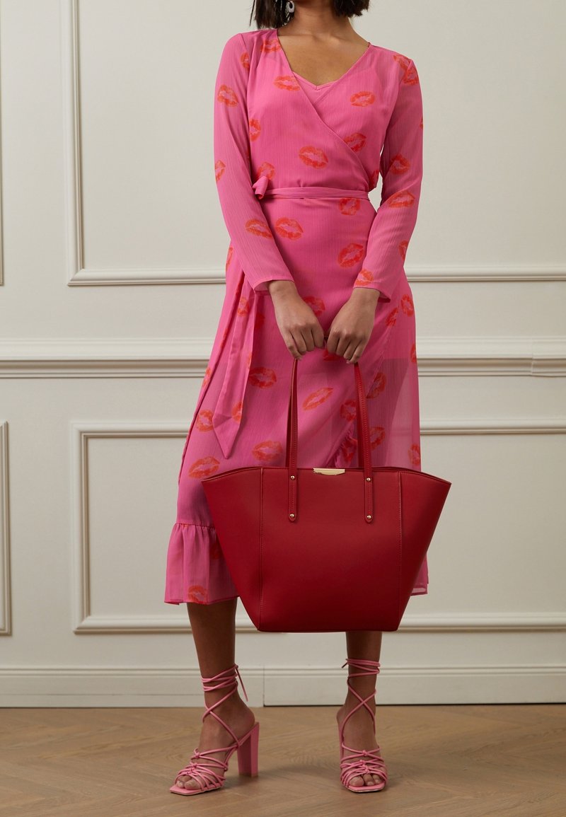 Femme en robe portefeuille rose avec des empreintes de lèvres rouges tenant un grand sac cabas rouge, portant des sandales roses à talons blocs dans un cadre intérieur.