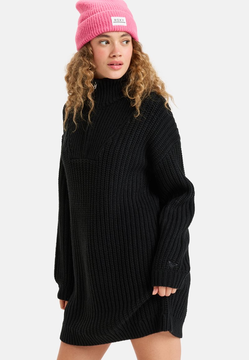 Pull en tricot noir surdimensionné avec de longues manches et un col montant, présentant un motif texturé et un petit logo sur la manche.
