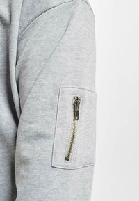 Der graue Sweatshirtstoff verfügt über eine Reißverschlusstasche am Ärmel. Die Textur ist glatt, und das Design ist lässig mit dezenten Details.