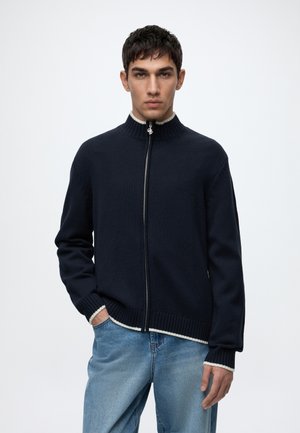 Giovane uomo che indossa un maglione blu navy con zip e bordi bianchi e jeans azzurri, in piedi con una mano in tasca contro uno sfondo semplice.