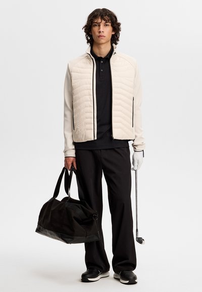 Ung mand i en beige quiltet jakke og sorte bukser, der holder en golfkølle og en sort duffeltaske mod en ensfarvet baggrund.
