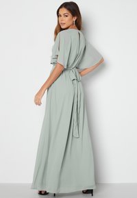 Langes, fließendes Maxikleid aus sanftem grünen Chiffon mit breiten Ärmeln, einem gebundenen Taillengürtel und einem Schlüsselloch-Detail im Rücken. Sanfte Textur mit dezenter Drapierung.