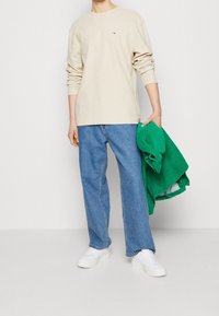 Beżowa koszula z długim rękawem z małym logo, w połączeniu z jasnoniebieskimi spodniami o szerokich nogawkach. Osoba trzyma zieloną kurtkę i ma na sobie białe trampki.