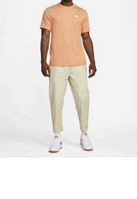 Persikofärgad Nike t-shirt med vit logotyp, tillsammans med ljusbeige tapered-byxor och vita sneakers med röda accenter. Avslappnad passform, mjukt tyg.