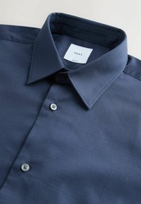 Marineblauw slim fit overhemd met een klassieke kraag en twee zichtbare knopen, met het witte merklabel "NEXT" zichtbaar aan de binnenkant van de kraag.