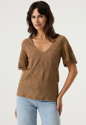 Vrouw draagt een bruin patroon gebreid kortärmelig shirt met een V-hals en lichtblauwe spijkerbroek, staand tegen een effen achtergrond.