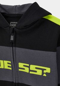 Schwarze und graue Zip-Hoodie mit neon-gelbem "GUESS?" Logo auf der Brust und einem neon-gelben Streifen an der Schulter.