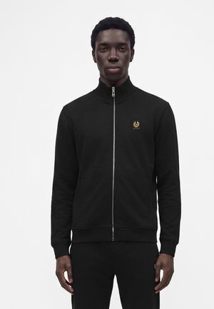 Junger Mann trägt eine schwarze Jacke mit Reißverschluss, hohem Kragen und kleinem goldenem Emblem auf der Brust, steht vor einem schlichten weißen Hintergrund.