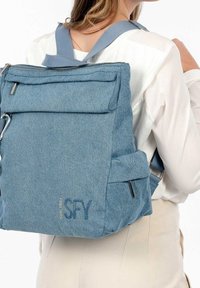 Blå denimryggsäck med justerbara remmar, frontficka med dragkedja och sidofickor. Utrustad med metallbeslag och tryckt text.