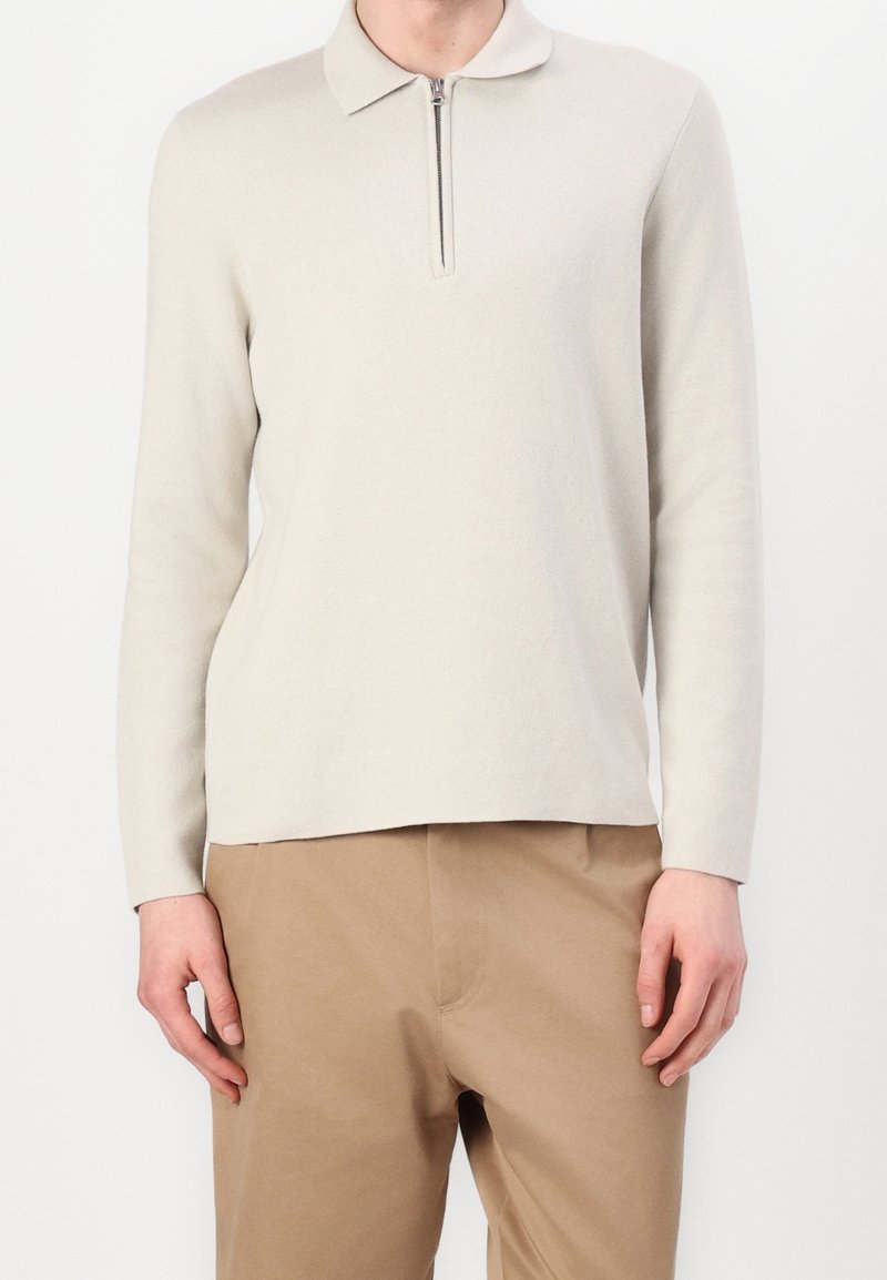 Homme portant un polo à manches longues beige clair avec une fermeture éclair à l'avant, associé à un pantalon marron clair sur un fond blanc uni.