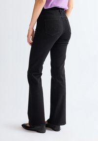 Frau trägt schwarze, hoch taillierte gerade Jeans mit Gesäßtaschen, kombiniert mit schwarzen Ballerinas und einem in die Hose gesteckten lila Oberteil.