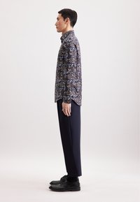 Bloemendessin shirt in navy met blauwe en beige accenten, gecombineerd met donkere broeken en zwarte schoenen. Gladde stof, op maat gesneden pasvorm. Zij-aanzicht weergegeven.
