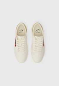 Krémovo-biele tenisky s červenými akcentmi, textilný zvršok, okrúhla špička a plochá gumová podrážka. Obsahuje logo na jazyku a šnurovanie.