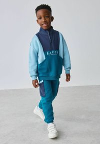 Chándal azul y verde azulado con cuello alto, que presenta un logo blanco y acentos de bolsillo en contraste. Llevado con zapatillas blancas.