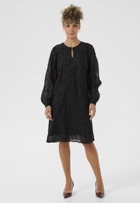 Femeie stand în picioare, îmbrăcată cu o rochie neagră până la genunchi, cu mâneci lungi transparente și broderie florală, asortată cu pantofi cu vârf ascuțit burgundy.