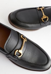 Mocassins en cuir noir avec une finition texturée, ornés d'un détail de quincaillerie dorée. Bout arrondi et coutures contrastantes le long des bords.