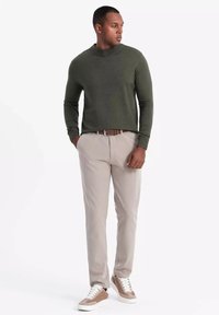Camicia a maniche lunghe verde oliva con collo alto, abbinata a pantaloni beige chiaro e sneakers con dettagli marroni. Il tessuto sembra morbido e strutturato.