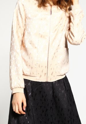 Frau trägt eine hellrosa Zip-up-Jacke mit einem dezenten geometrischen Muster und einen schwarzen Rock mit ähnlicher geometrischer Struktur.