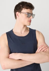 Marineblå, ermeløs bomullstopp med subtil brodert "BOSS"-logo. Modell med sølvfarvede solbriller, med armene krysset.