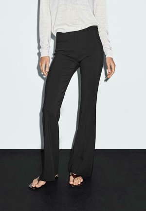 Zwarte uitlopende broek met een gladde textuur, hoge tailleband en subtiele zijzakdetails, gecombineerd met een lichtgrijze lange mouwtop.