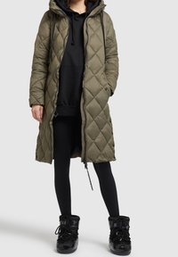 Olive green quilted long coat with a zipper and snap buttons, featuring a hood and ties. Worn over a black sweatshirt and leggings, paired with black boots.