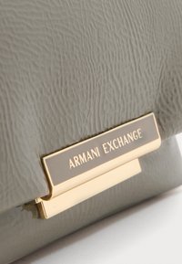 Artimi nuotrauka su tekstūruota pilka odine rankine, turinčia auksinės spalvos metalinį užsegimą su išgraviruotu „ARMANI EXCHANGE“ logotipu.