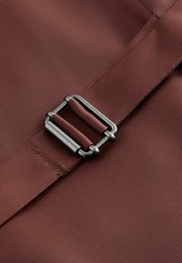 Sangle ajustable en tissu bordeaux passée à travers une boucle métallique rectangulaire sur un fond textile bordeaux assorti.
