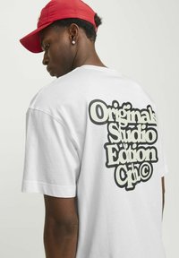 Valge puuvillane t-särk, mille seljal on suur must graafiline tekst, mis ütleb "Originals Studio Edition Cph", lühikeste varrukatega.