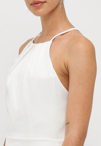 White halter neck dress with pleated front detail, smooth texture, and thin straps. Close-up view of shoulder and upper chest area.