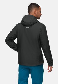 Uomo che indossa un giubbotto con cappuccio nero con il logo "Mammut" sulla schiena e pantaloni blu brillanti, rivolto di spalle su uno sfondo semplice.