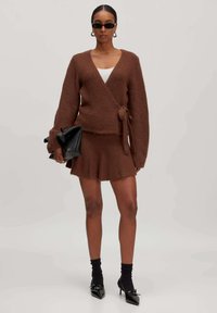 Brauner, flauschiger Wickelpullover mit langen Ärmeln und Bindedetail, kombiniert mit einem passenden, ausgestellten Minirock. Zubehör umfasst eine schwarze Tasche und Sonnenbrille.