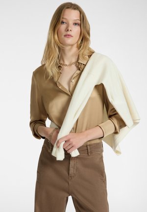 Camicia di satin marrone chiaro con dettagli a bottone, abbinata a pantaloni marroni. Un maglione lavorato a maglia color crema è appoggiato su un braccio. Bracciale a catena visibile.