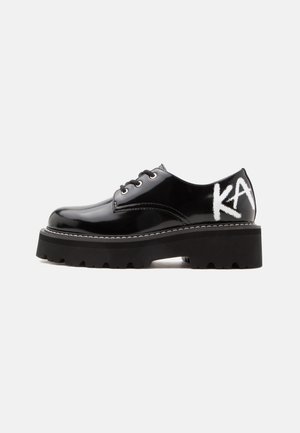 Chaussure noire brillante à lacets avec semelle épaisse et texte blanc "KA" sur le talon, vue de côté sur fond blanc.