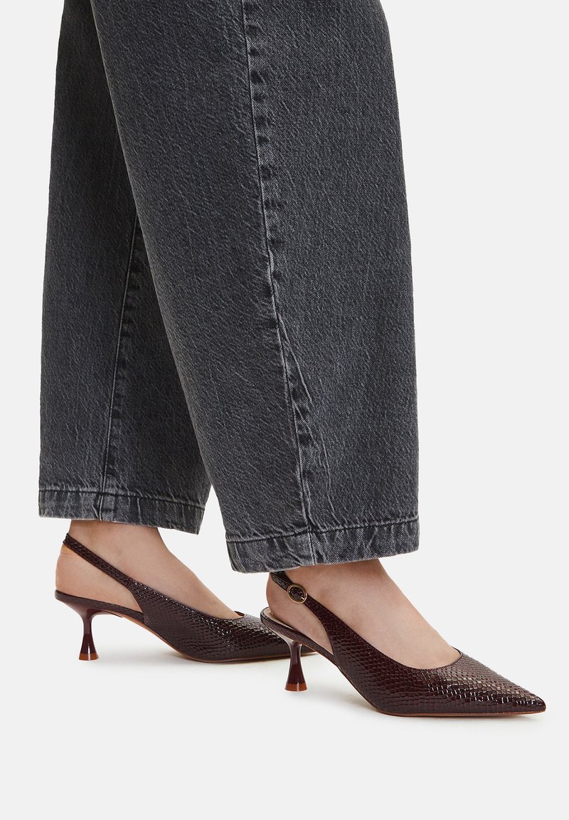 Brązowe, skórzane buty na obcasie typu slingback z wyraźną teksturą, mające spiczasty nos i mały, zakrzywiony obcas, noszone z szerokimi szarymi spodniami.