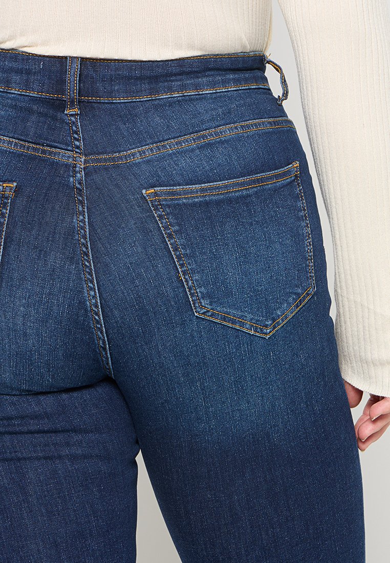 Dunkelblaue Jeans aus Denim mit hohem Bund, Gesäßtaschen und orangefarbenen Nähten. Der Stoff hat eine glatte, leicht strukturierte Optik.
