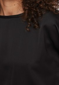 Haut noir en tissu lisse, avec un col rond et une coupe décontractée. Des cheveux bouclés sont visibles en arrière-plan.