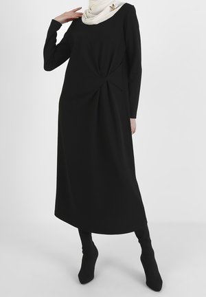 Kvinde iført en sort midi-kjole med lange ærmer og løgdetalje ved taljen, kombineret med sorte støvler og et hvidt tørklæde med broche.